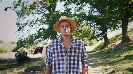 Young villager man shepherd in straw hat walking with his flock of cows on a rural background, slow motion