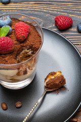 spoon with piece of Classic tiramisu dessert with blueberries and strawberries in a glass cup on wooden background