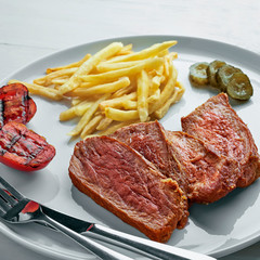 Medium rare sirloin steak with chips, pickled cucumber, grilled cherry  and a glass of sparkling water on a white wooden table with tablecloth. Classic European lunch.