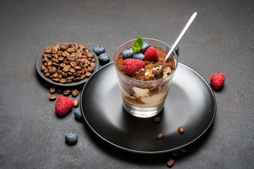 Classic tiramisu dessert portion with blueberries and raspberries in a glass cup on stone serving board on concrete background