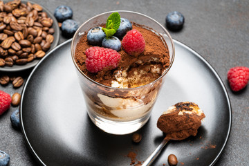 Classic tiramisu dessert portion with blueberries and raspberries in a glass cup on stone serving board on concrete background