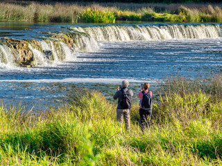 Travelers photographed on smartphones beautiful waterfall. Rumba is a waterfall on the Venta River...
