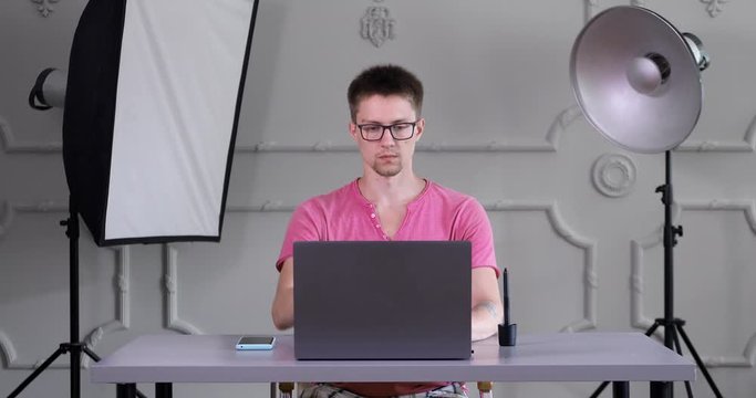 a guy with glasses and a pink shirt sits at a table and works at a laptop in an interior photo studio