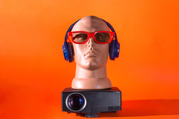 mannequin in blue headphones and red 3D glasses with a projector for home theater on orange isolated background