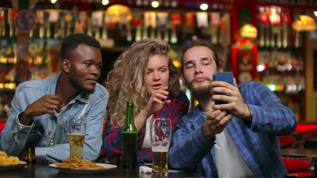 Multi-ethnic group of friends take a selfie in a bar and laugh with a beer while looking at photos on a smartphone screen.