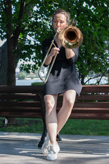 Girl learning to play trombone. Girl plays leaning on a park bench.