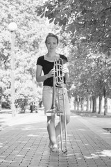 Girl learning to play trombone. Girl plays standing on the alley of a city park.