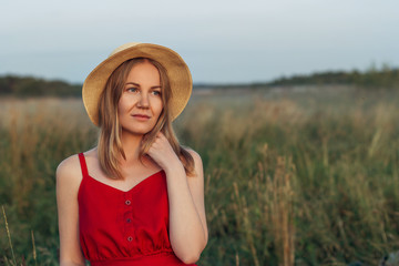 portrait of a young beautiful girl in a red dress and a straw hat at sunset, she is thoughtful, look away