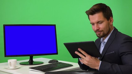 A young businessman sits in front of a computer with blue screen and works on a tablet, then smiles at the camera - green screen studio