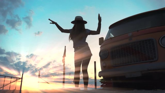 Retro style transport concept. Sexy girl dancing in amazing sun light at the sea beach near the vintage car. Bright sun rays illuminating body movements. Retro van in sunset. Positive tourist enjoying