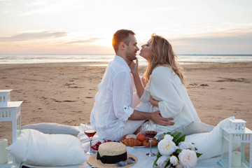 Picnic at sunset. Man and a pregnant woman sit on a bedspread, on the beach, hugging, kissing against low tide.