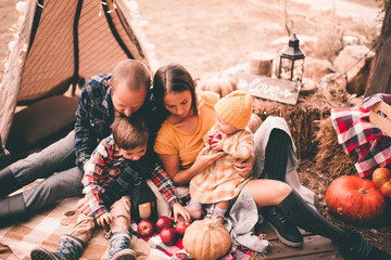 Family having rest outdoors with autumn pumpkins and apples. Harvesting time. Mother, father and children wearing trendy autumn clothes having picnic.