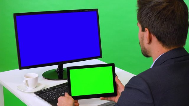 A young businessman sits at a desk with a blue computer screen and looks at a tablet with green screen - closeup - green screen studio