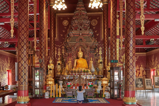 nside a Buddhist temple in Thailand, chaired by a large golden Buddha, in the middle there is a tourist on her knees who takes pictures