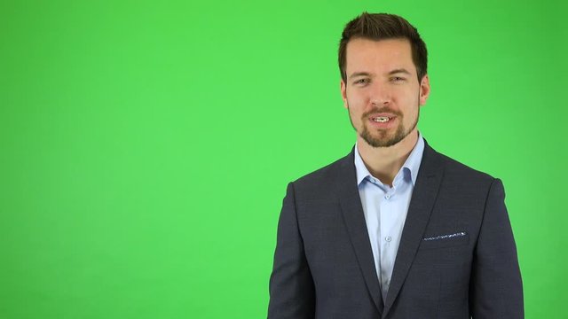 A young handsome businessman talks to the camera with a smile, gestures toward it and next to himself - green screen studio