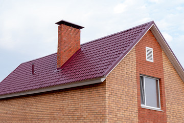 Part of a two-story private building of red brick against a clear sky. Fragment of a roof with a pipe and a window on the house. New affordable housing under the state program or mortgage lending.