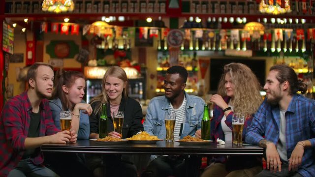 African American with friends at the bar drinks beer and eats chips, friends raise glasses and bottles and knock / check on the table smiling and saying toasts