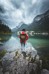 People and nature concept. Female tourist enjoys morning view from shore, looks at mountains and lake, being solo traveller, watching beauty, carries backpack, dressed in red raincoat and a hat.