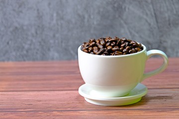 White coffee cup filled up with roasted coffee beans placed on wooden table with concrete background.