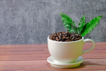 White coffee cup filled up with roasted coffee beans with smoke decorated with green coffee leaves placed on wooden table with concrete background.