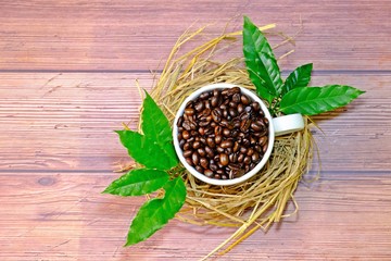 Top view coffee beans in white coffee cup placed on small nest decorated with green coffee leaves on wooden table.