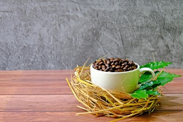 Coffee beans in white coffee cup placed on small nest decorated with green coffee leaves on wooden table.