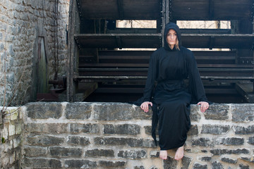 Young Woman with dark long hair in black robes in front of an Old Wooden Water Mill. Witches. Halloween concept. Witchcraft and Magic. 