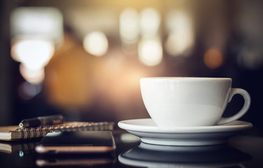 Closeup white cup of coffee with smartphone, notebook and pen on table in cafe. Vintage light, blurred and bokeh background - Powered by Adobe