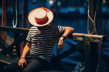 Gondolier in  hat and striped clothes sits in  evening on canal and waits for client with mobile phone - obrazy, fototapety, plakaty