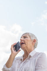 older woman with White shirt and White hair talking with mobile, sky background