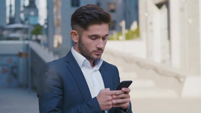 Young Handsome Businessman Standing near Big Office Building. Typing a Message on his Smartphone.Bearded Man Wearing Classical Suit. Business Lifestyle.