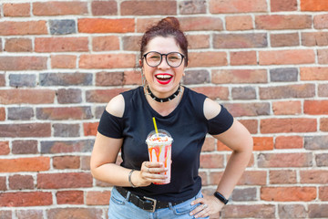 Fototapeta premium Cute cheerful young woman in stylish clothes is laughing and holding a milkshake in her hands against a brick wall. The concept of a summer holiday in the city. Copyspace.