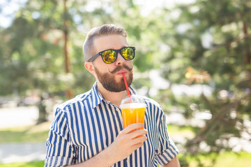 Fototapeta premium Portrait of a positive cheerful young man with a glass of juice with a straw while walking in the park on a warm sunny summer day. The concept of rest after study and work on weekends.