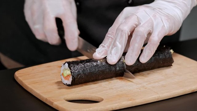 Young male sushi chef prepares Japanese sushi rolls of rice, salmon, avocado and nori. Restaurant kitchen, closeup hands cut a roll with a knife.