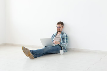 Positive young man in casual clothes surfing the Internet in search of new housing sitting on the floor in an empty room. The concept of finding an apartment using the Internet and a laptop.