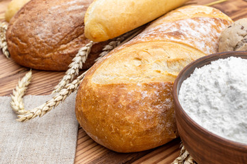 Loafs of bread with flour and wheat ears on wooden background.