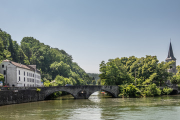 Fototapeta premium Dinant, Belgium - June 26, 2019: Old roman bridge over greenish Lesse River. Saint Anne Church tower captured in green foliage on the right. White building on the left under light blue sky.