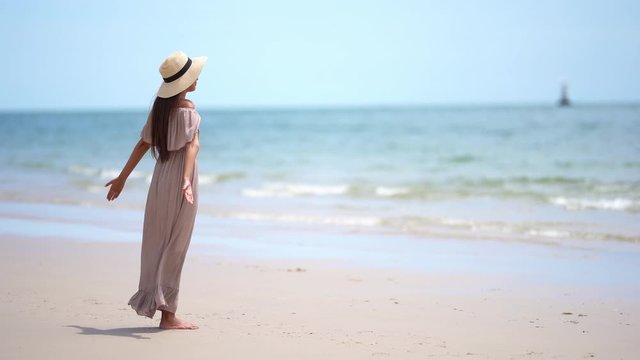 Young Asian woman in a pink flowing sundress and floppy sun hat stands on a windy beach arms stretch back enjoying the waves and ocean breeze.