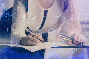 A woman hands writing information about stock market in notepad. Forex chart holograms in front....