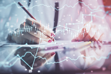 A woman hands writing information about stock market in notepad. Forex chart holograms in front. Concept of research.