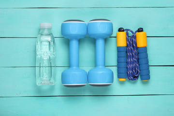 Flat lay style sport concept. Dumbbells, jump rope, bottle of water. Sports equipment on blue wooden background. Top view