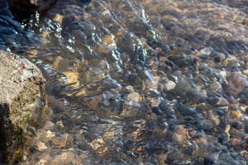 Klares Felsquellwasser fließt von der Quelle in einen Bach und später in einen klaren Fluss mit schöner Strömung, Reflexionen von Sonnenlicht und Spiegelungen und zeigt schöne Kieselsteine am Grund