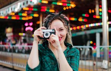 An informal young woman with dreadlocks takes pictures with retro camera in amusement park.