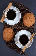 two white coffee mugs on a saucer on a stone table with scattered coffee beans, oatmeal cookies chocolate and cinnamon stick, top view