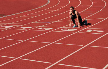Young runner woman in sportwear getting ready to run sprint at low start on stadium track with red...