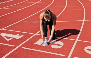 Young sprinter woman in sportswear tying shoelaces before a race on a red-coated stadium track at sunny bright day