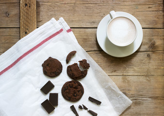 Chocolate cookies, pieces of chocolate and a cup of cocoa or coffee with milk on a wooden background. Rustic style.