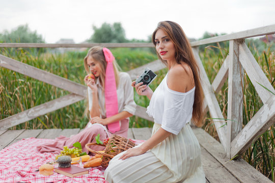 Two cheerful young women have a picnic outdoors on a summer day. Two girlfriends in retro vintage style clothes spend time together and taking photo with retro camera on the pond pier.