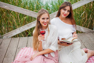 Two cheerful young women have a picnic outdoors on a summer day. Two girlfriends in retro vintage style clothes spend time together on the pond pier.