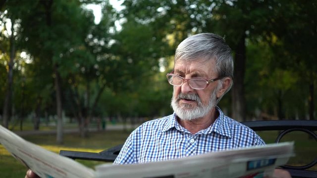 senior in plaid shirt sits on a bench in the park and reads a newspaper